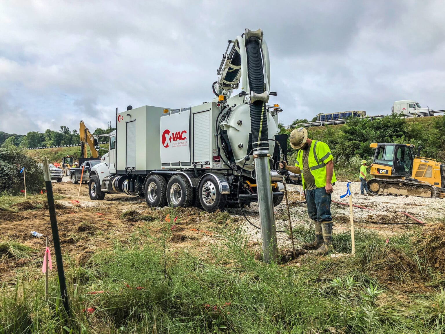 White X-Vac hydro excavation truck with boom and large suction hose at a roadside worksite, worker nearby
