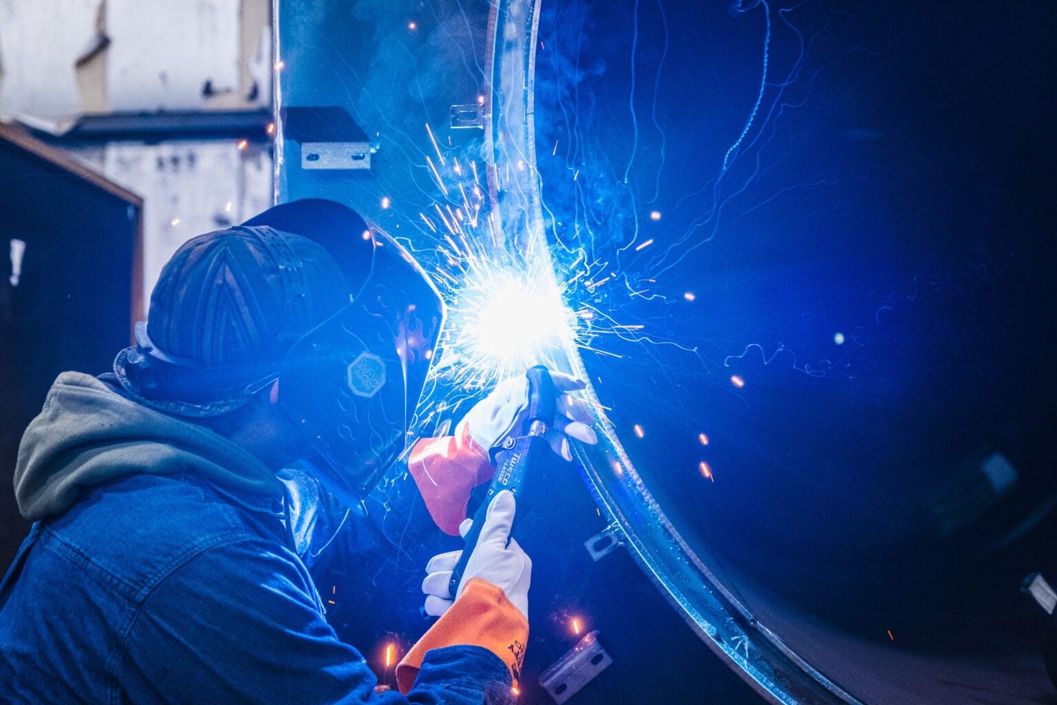 Welder in helmet and gloves welding a curved metal panel with blue sparks, illustrating X-Vac American manufacturing.