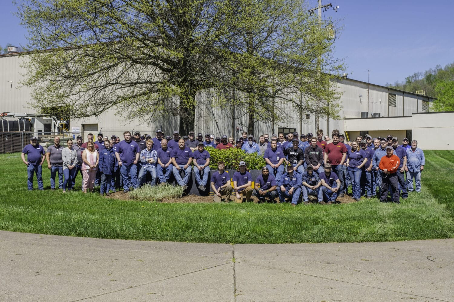 Large X-Vac hydro excavation crew posed in front of their manufacturing building under a blossoming tree.