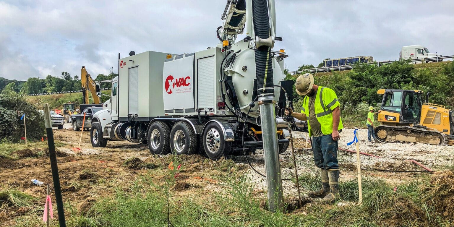 White X-Vac hydro excavation truck with boom and large suction hose at a roadside worksite, worker nearby