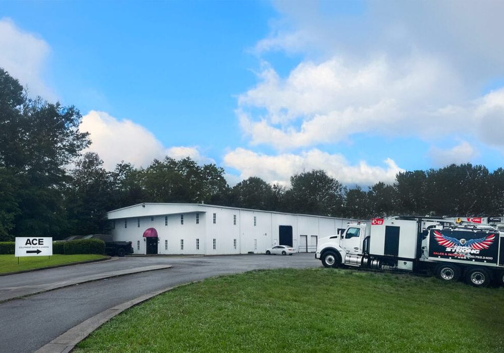 White industrial building and treeline under blue sky; X-Vac hydro-excavation truck parked right, ACE sign