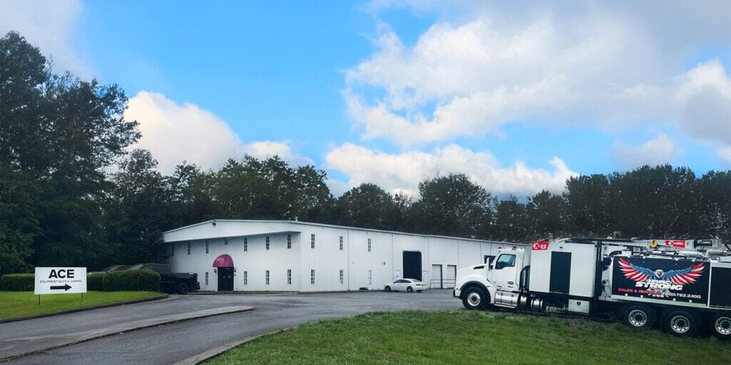 White industrial building and treeline under blue sky; X-Vac hydro-excavation truck parked right, ACE sign