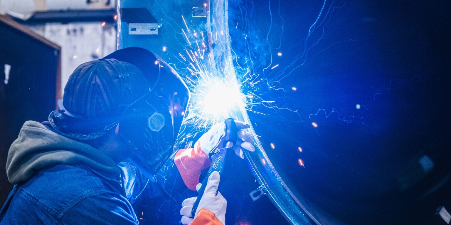 Welder in helmet and gloves welding a curved metal panel with blue sparks, illustrating X-Vac American manufacturing.